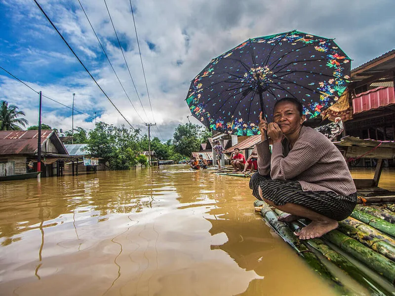 Seorang perempuan memegang payung menunggu dievakuasi tim penyelamat di atas rakit bambu di depan rumahnya yang terendam banjir di Desa Sungai Raya, Kabupaten Banjar, Kalimantan Selatan. (Foto: Putra/Greenpeace)