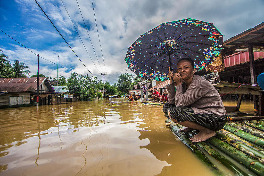 Seorang perempuan memegang payung menunggu dievakuasi tim penyelamat di atas rakit bambu di depan rumahnya yang terendam banjir di Desa Sungai Raya, Kabupaten Banjar, Kalimantan Selatan. (Foto: Putra/Greenpeace)